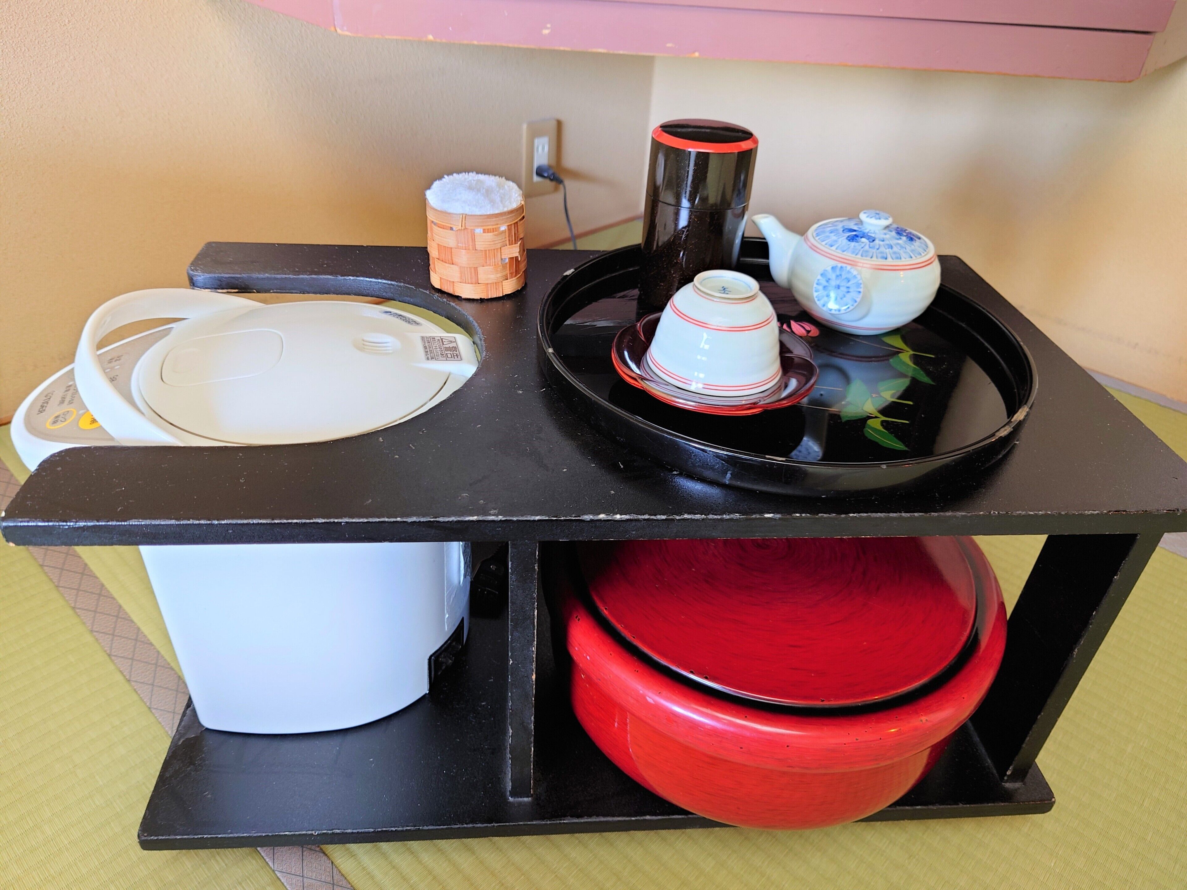 A kitchen counter with a rice cooker, a black thermos, a teapot, and stacked plates.