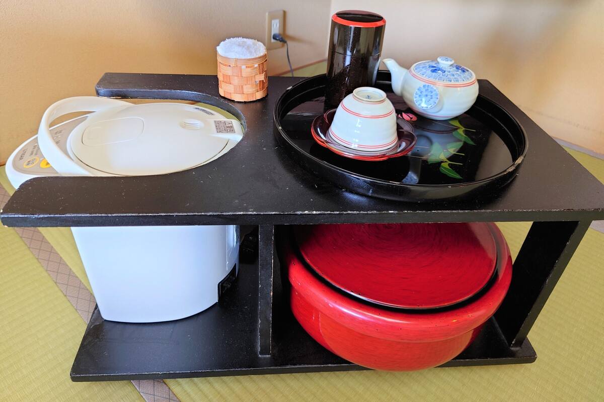 A kitchen counter with a rice cooker, a black thermos, a teapot, and stacked plates.