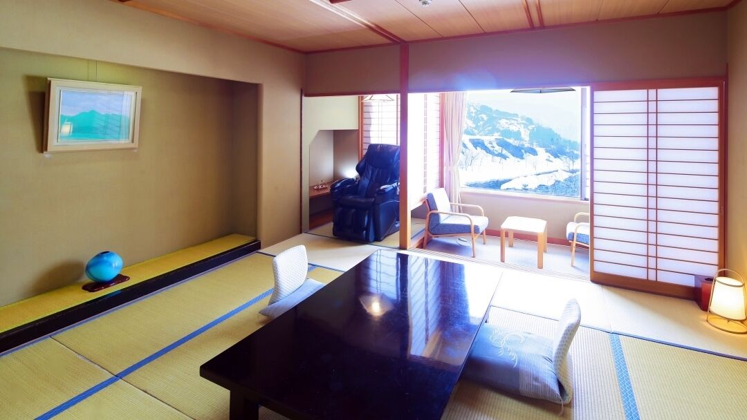 Una habitación con esteras de tatami, una mesa negra, un sillón de masaje y vistas a una montaña nevada.