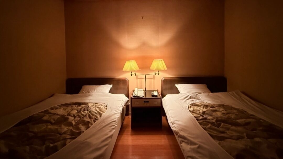 A hotel room with two single beds, a nightstand with a phone and lamp, and a wall-mounted lamp between the beds.