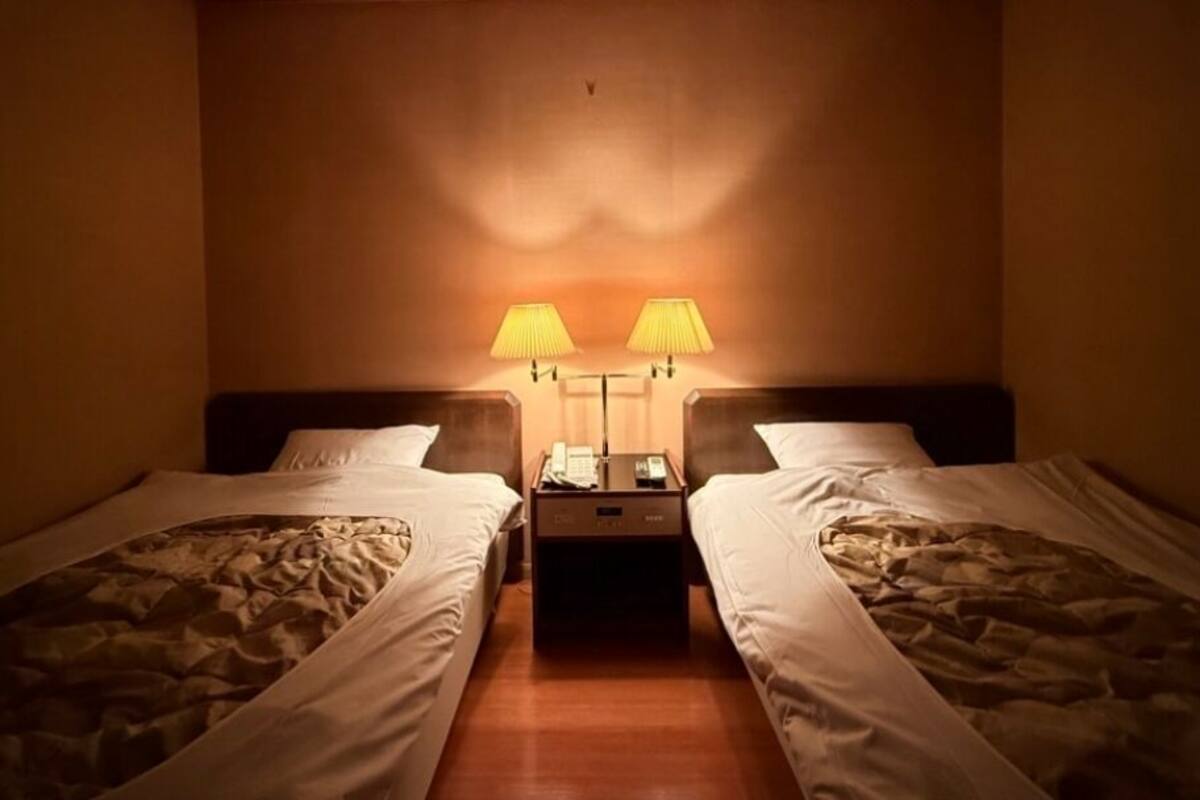 A hotel room with two single beds, a nightstand with a phone and lamp, and a wall-mounted lamp between the beds.