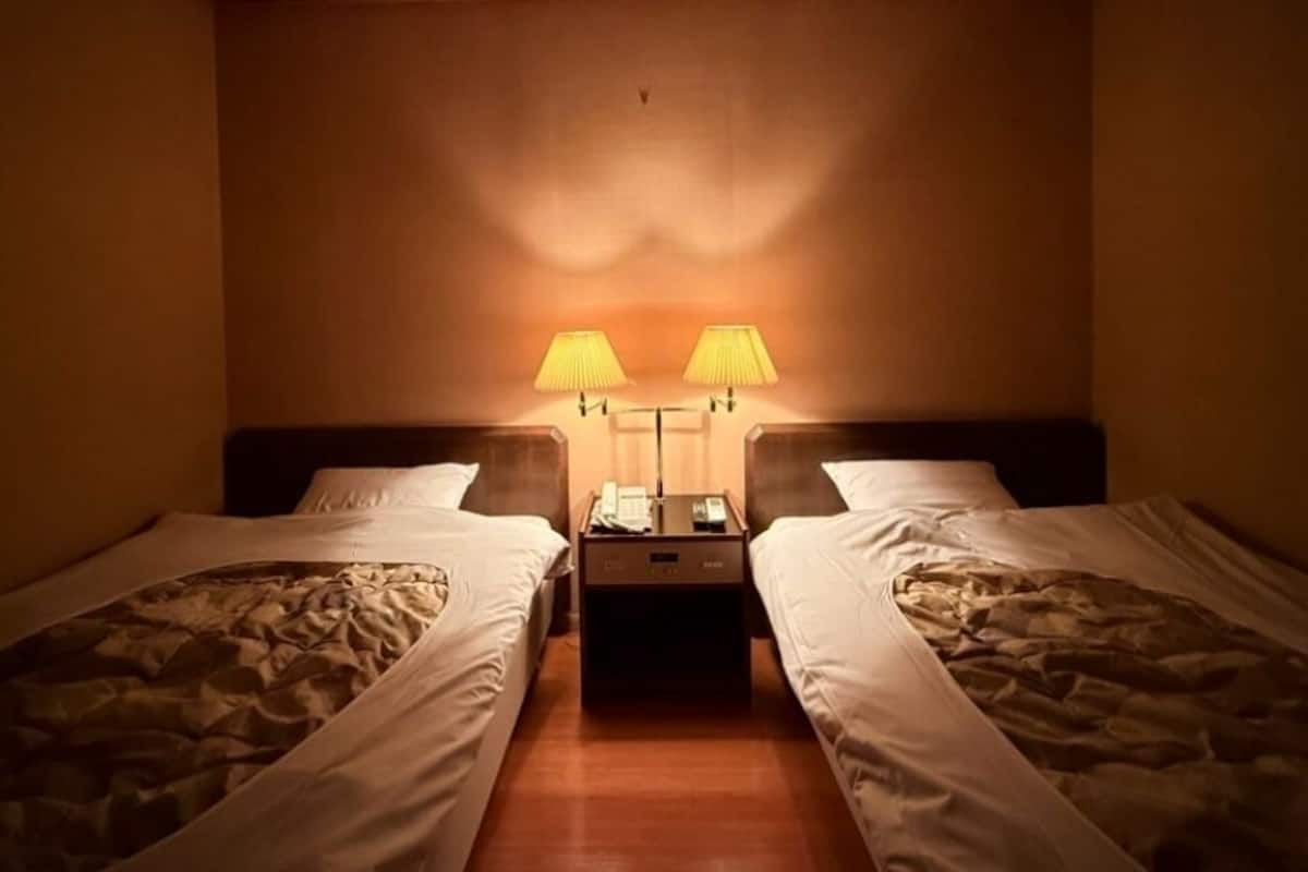 Hotellihuone, jossa on kaksi erillistä sänkyä, yöpöytä, jossa on puhelin ja lamppu, sekä seinään kiinnitetty lamppu sänkyjen välissä.
