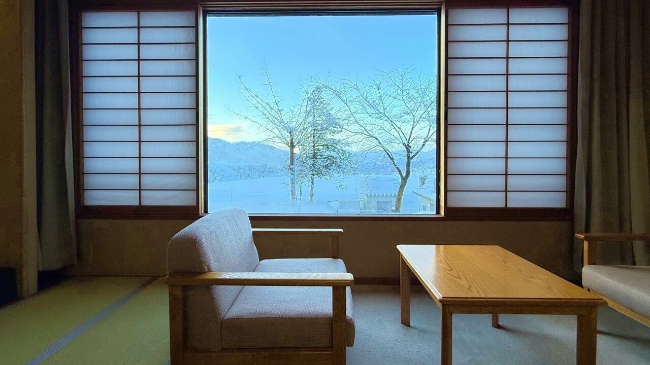 A room with tatami flooring, a low wooden table, a cushioned seating area, and a large window with shoji screens.