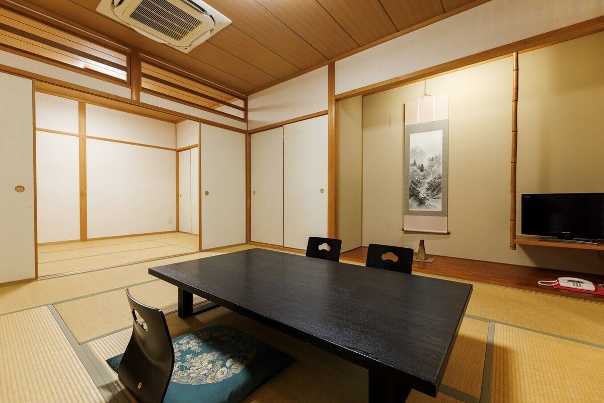 A traditional Japanese room with tatami flooring, a low table, chairs, a television, and a wall-mounted phone.