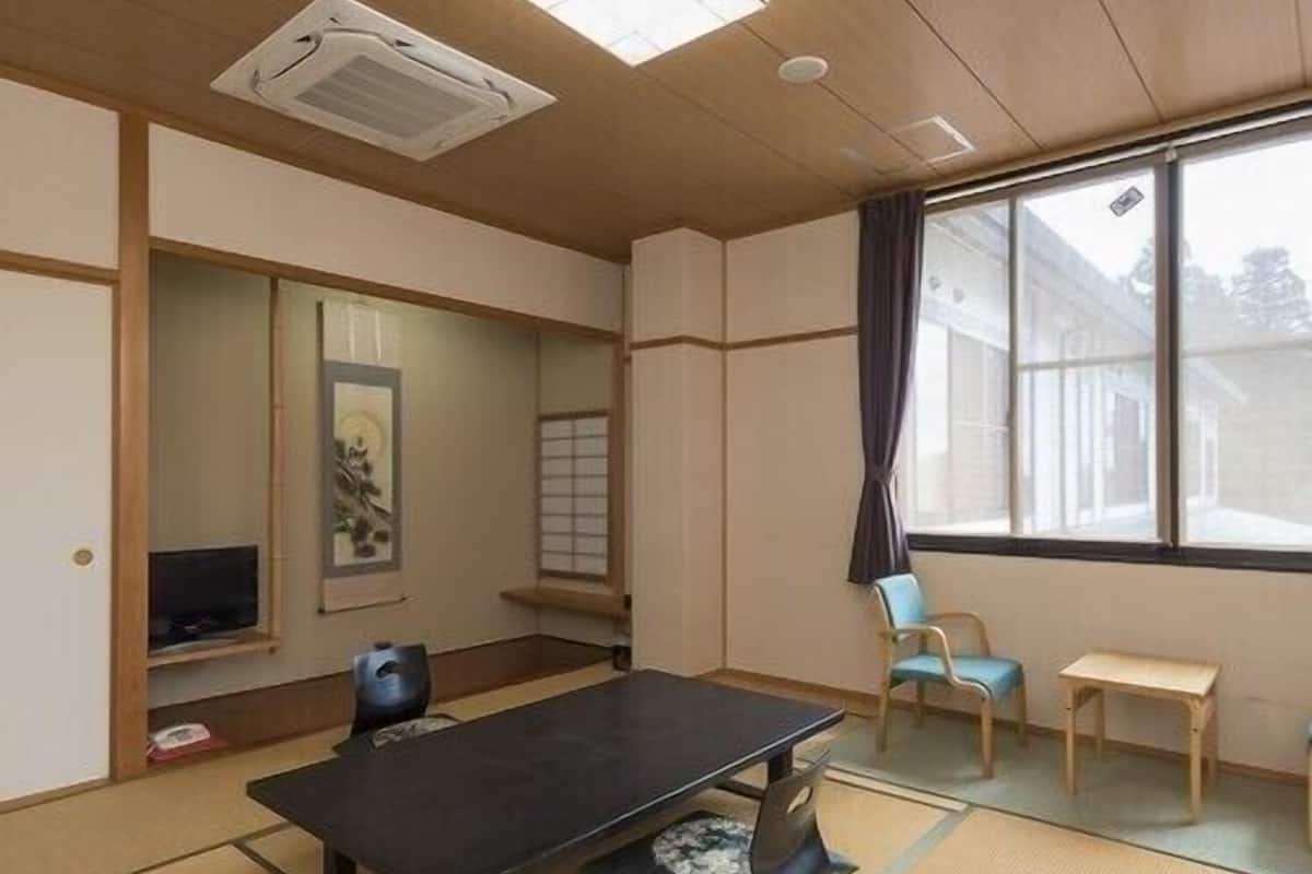 Ein traditioneller japanischer Raum mit Tatami-Bodenbelag, einem Tisch aus Tatami-Matten, einem Fernseher, einem Fenster mit Vorhängen und einer Schiebetür.