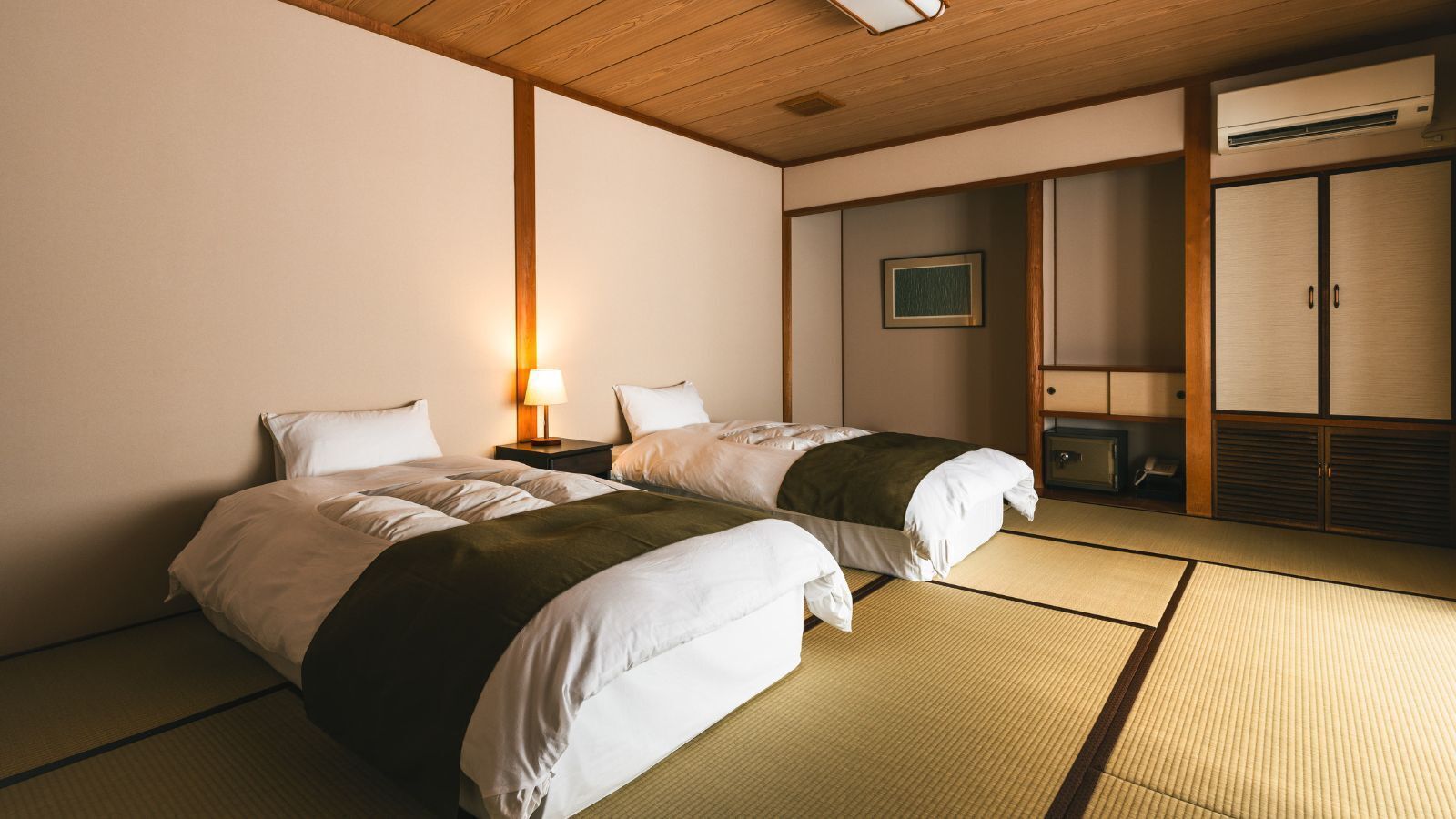A hotel room with two beds, a bedside table with a lamp, a wardrobe, and a framed picture on the wall.