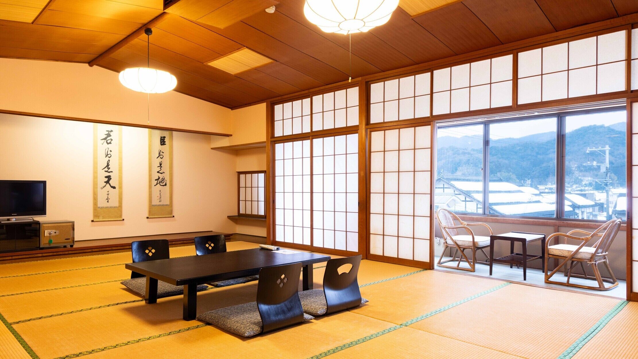 Ein traditioneller japanischer Raum mit Tatami-Böden, einem niedrigen Tisch mit Kissen, einem Fernseher und großen Fenstern mit Blick auf eine verschneite Landschaft.