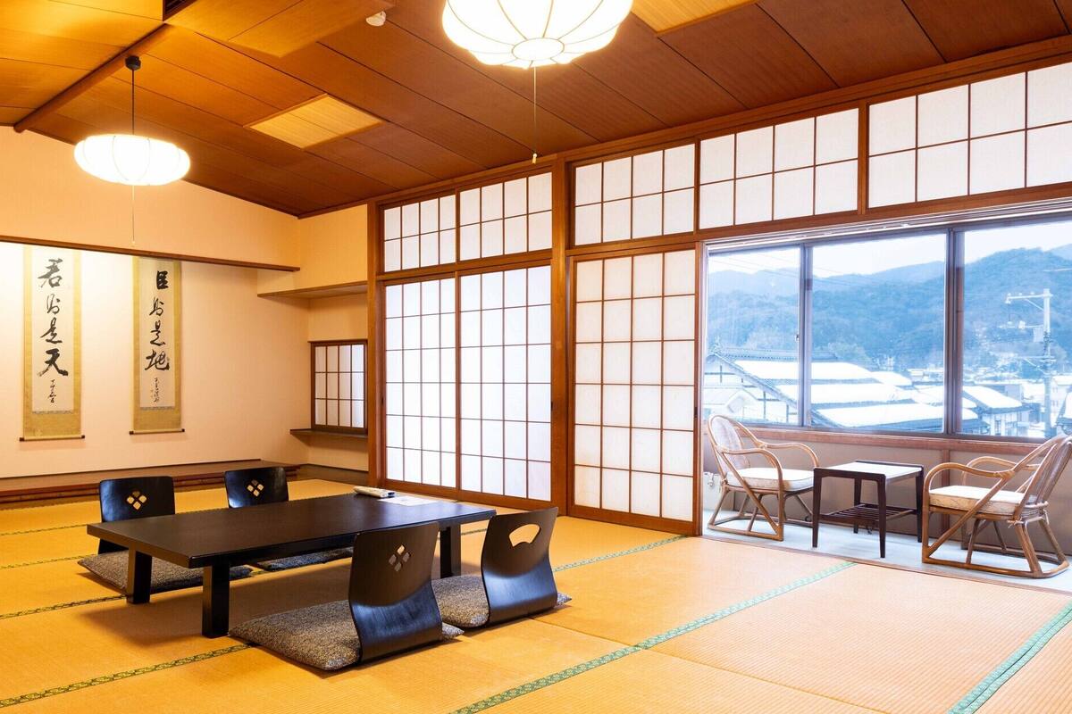 Una habitación japonesa tradicional con suelo de tatami, una mesa baja con cojines, un televisor y ventanas amplias con vistas a un paisaje nevado.