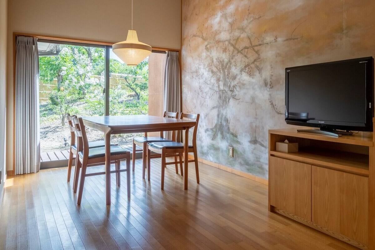 A room with a wooden dining table, chairs, a television on a wooden stand, and a view of greenery through a large window.