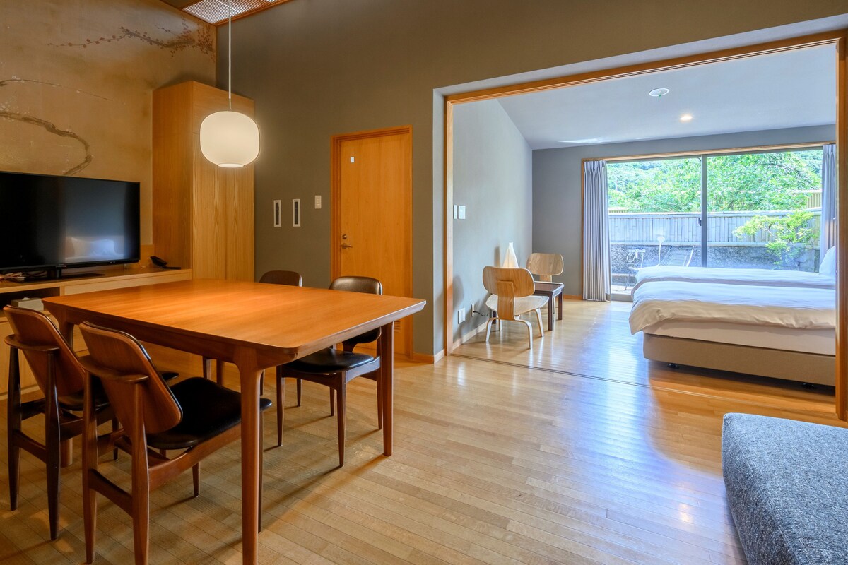 A modern hotel room with a wooden dining table, chairs, a TV, a bed, and a large window with a view of greenery.