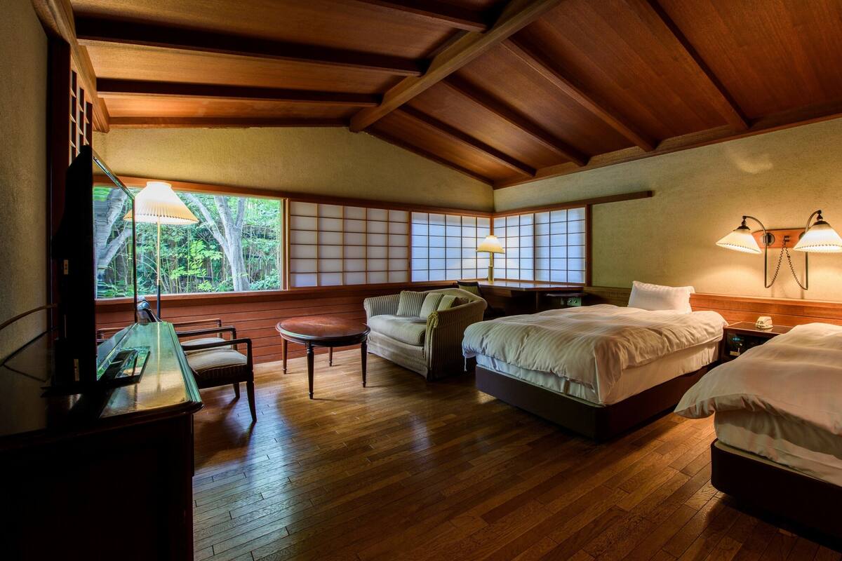 A hotel room with wooden flooring, two beds, a sofa, a table, and a view of greenery through large windows.