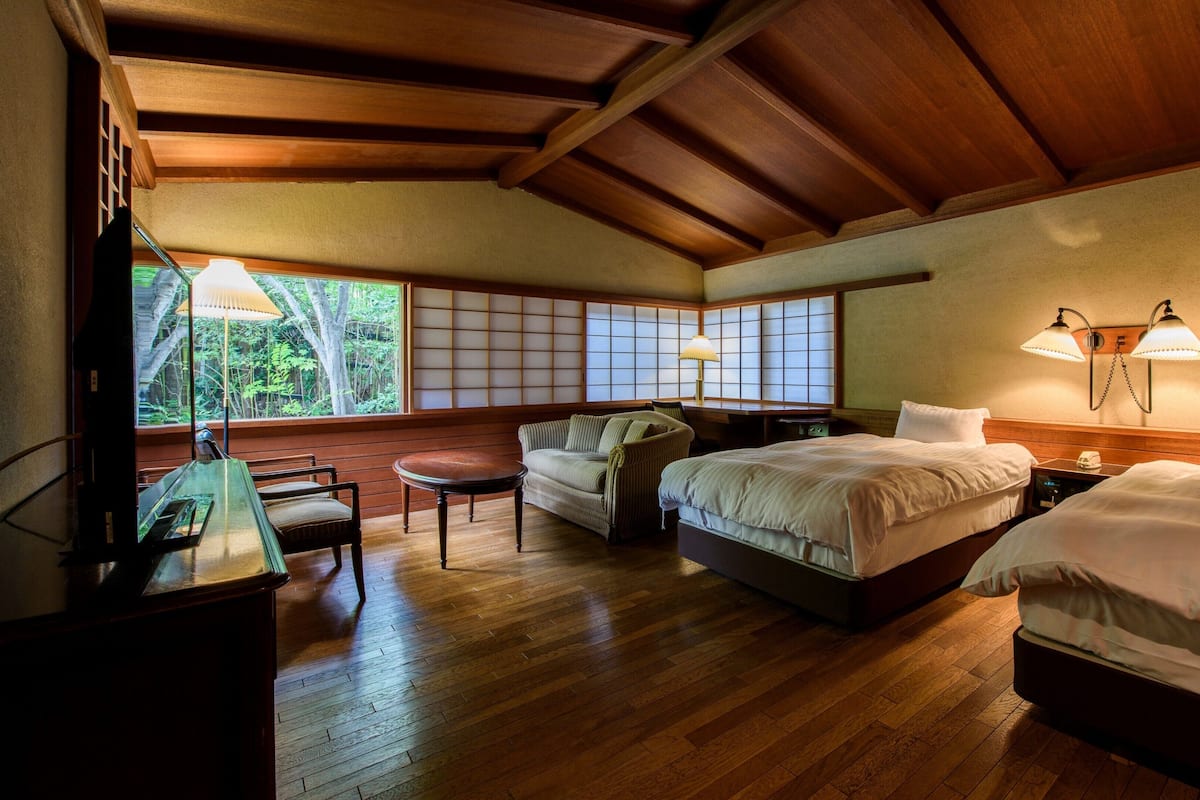 A hotel room with wooden flooring, two beds, a sofa, a table, and a view of greenery through large windows.