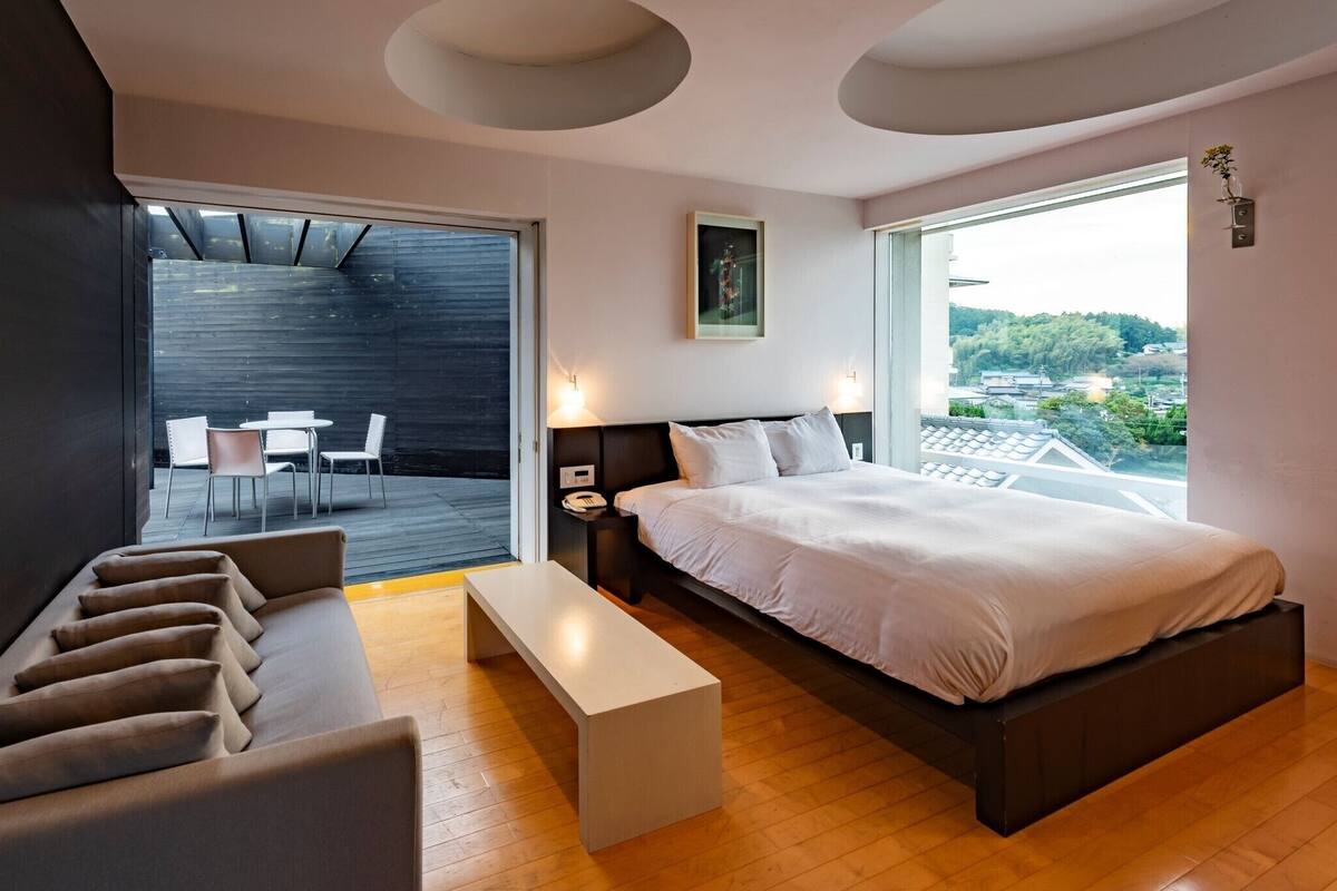 A modern bedroom with a large bed, a sofa, a small table, and a balcony with outdoor seating.