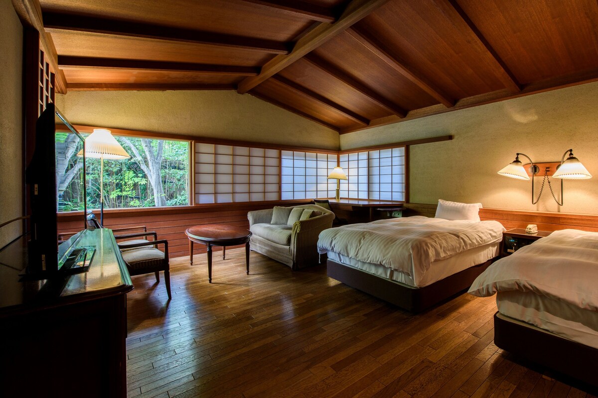 Habitación de hotel con suelo de madera, dos camas, un sofá, una mesa y vistas a zonas verdes a través de amplios ventanales.