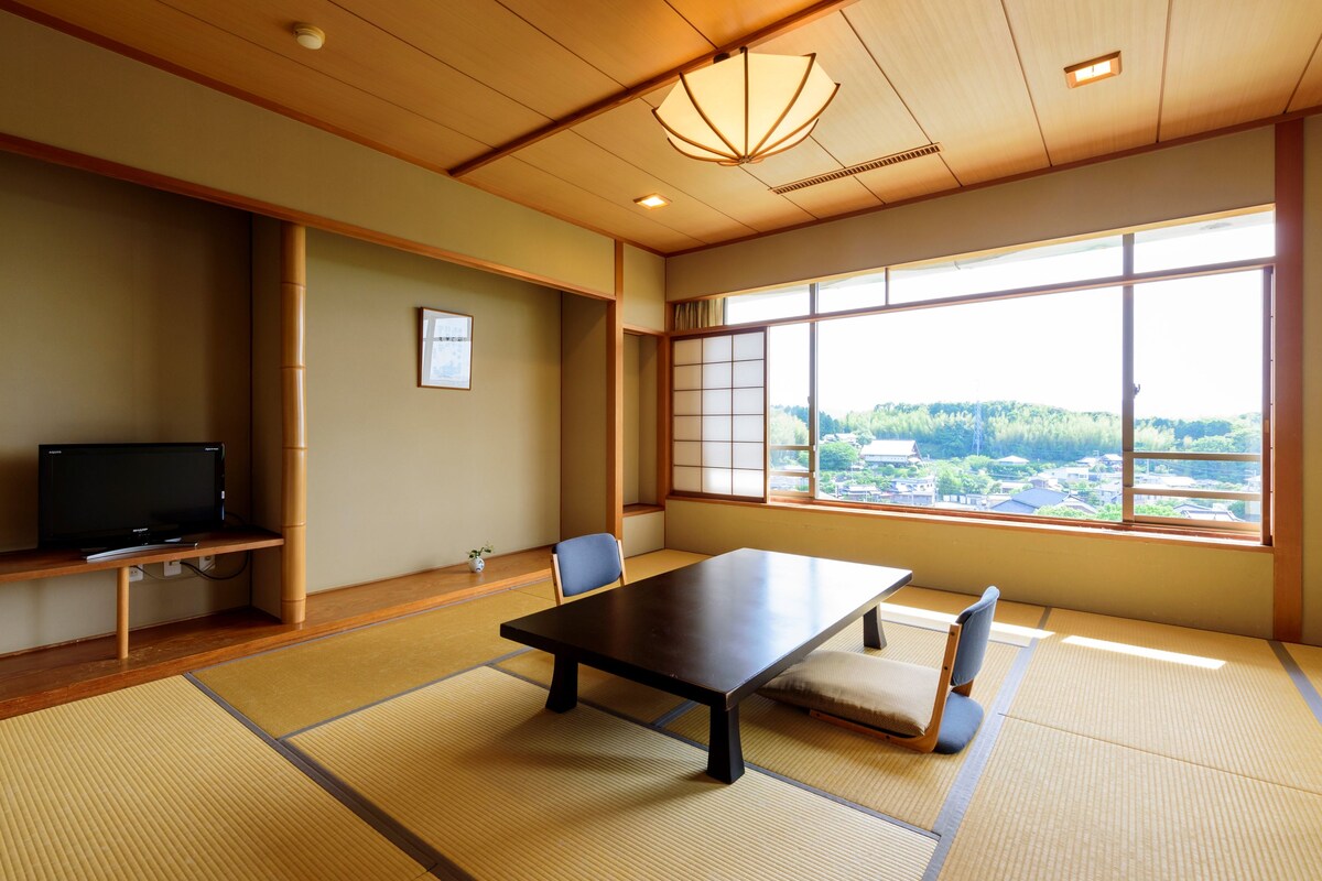 Une pièce de style japonais traditionnel, avec un sol recouvert de tatamis, une table basse, un téléviseur et une grande fenêtre donnant sur la ville.