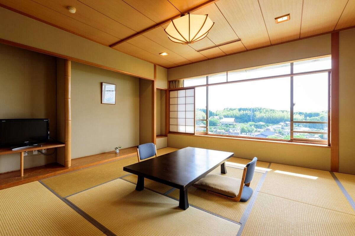 A traditional Japanese room with tatami flooring, a low table, a television on a shelf, and a large window with a view of a town.