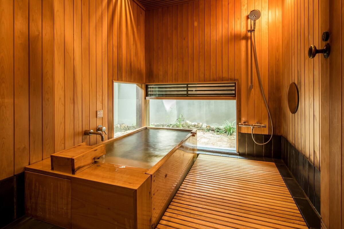 A wooden sauna room with a bench, a shower, and a view of greenery outside.