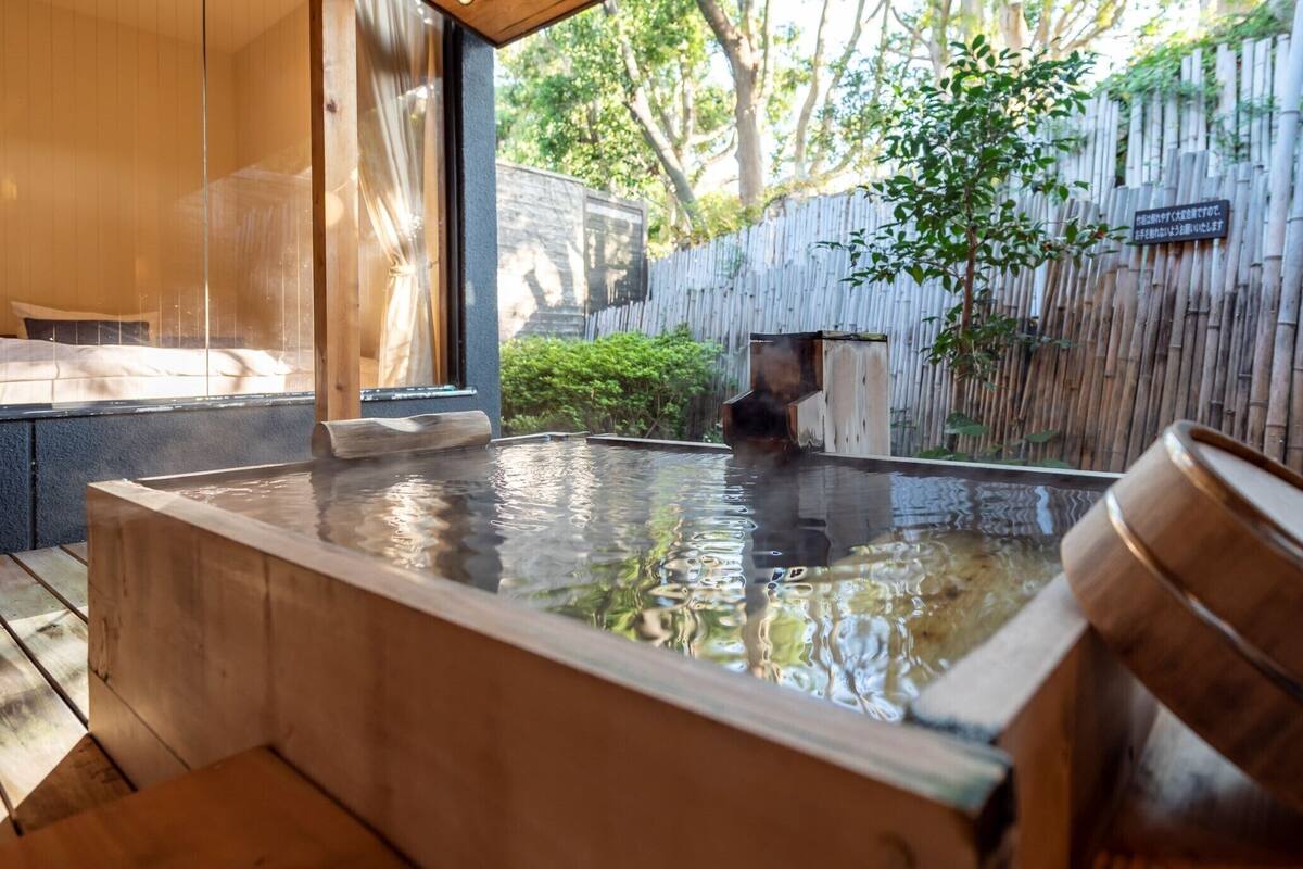A hot spring bath area with a wooden bench, a bamboo fence, and a view of the surrounding greenery.