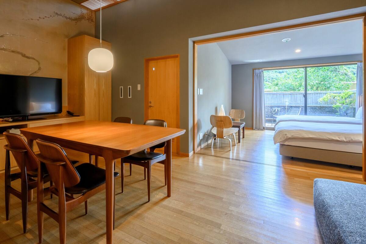 A modern hotel room with a wooden dining table, chairs, a TV, a bed, and a large window with a view of greenery.