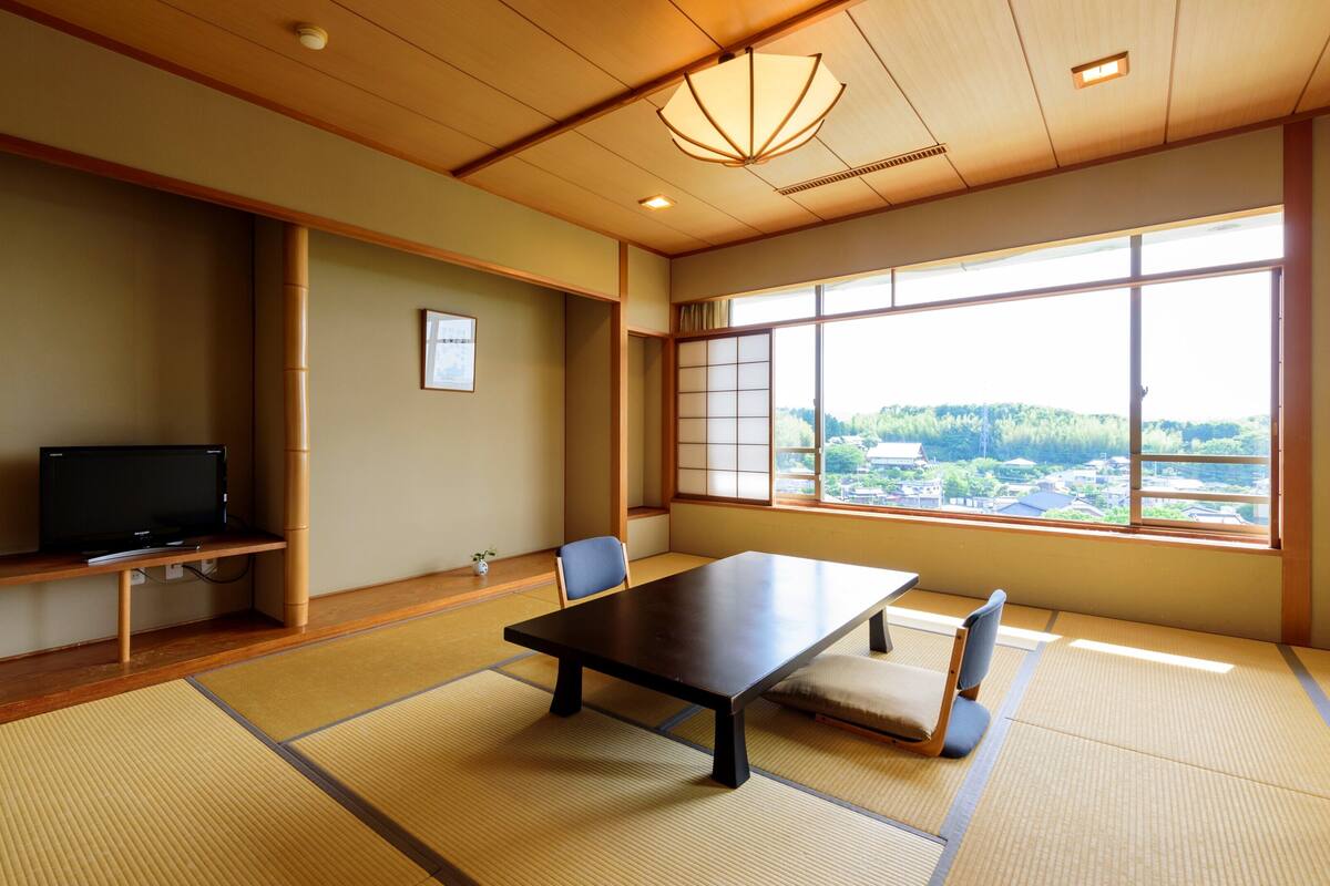 A traditional Japanese room with tatami flooring, a low table, a television, and a large window with a view of a town.