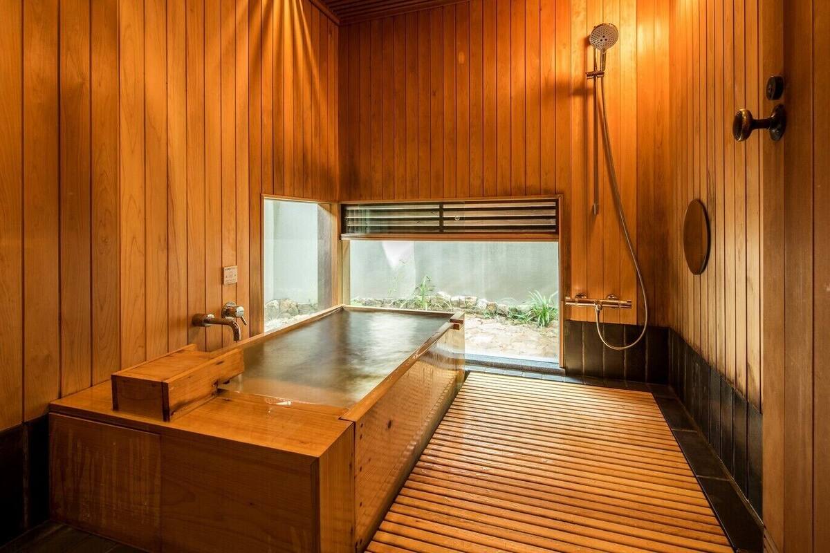 A wooden spa room with a large wooden bathtub, a shower area, and a view of a garden through a sliding door.