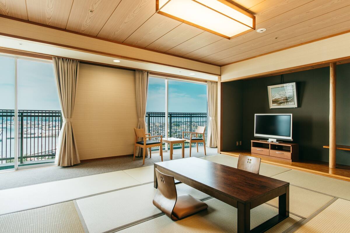A modern hotel room with a large window, a dining table, and a TV.