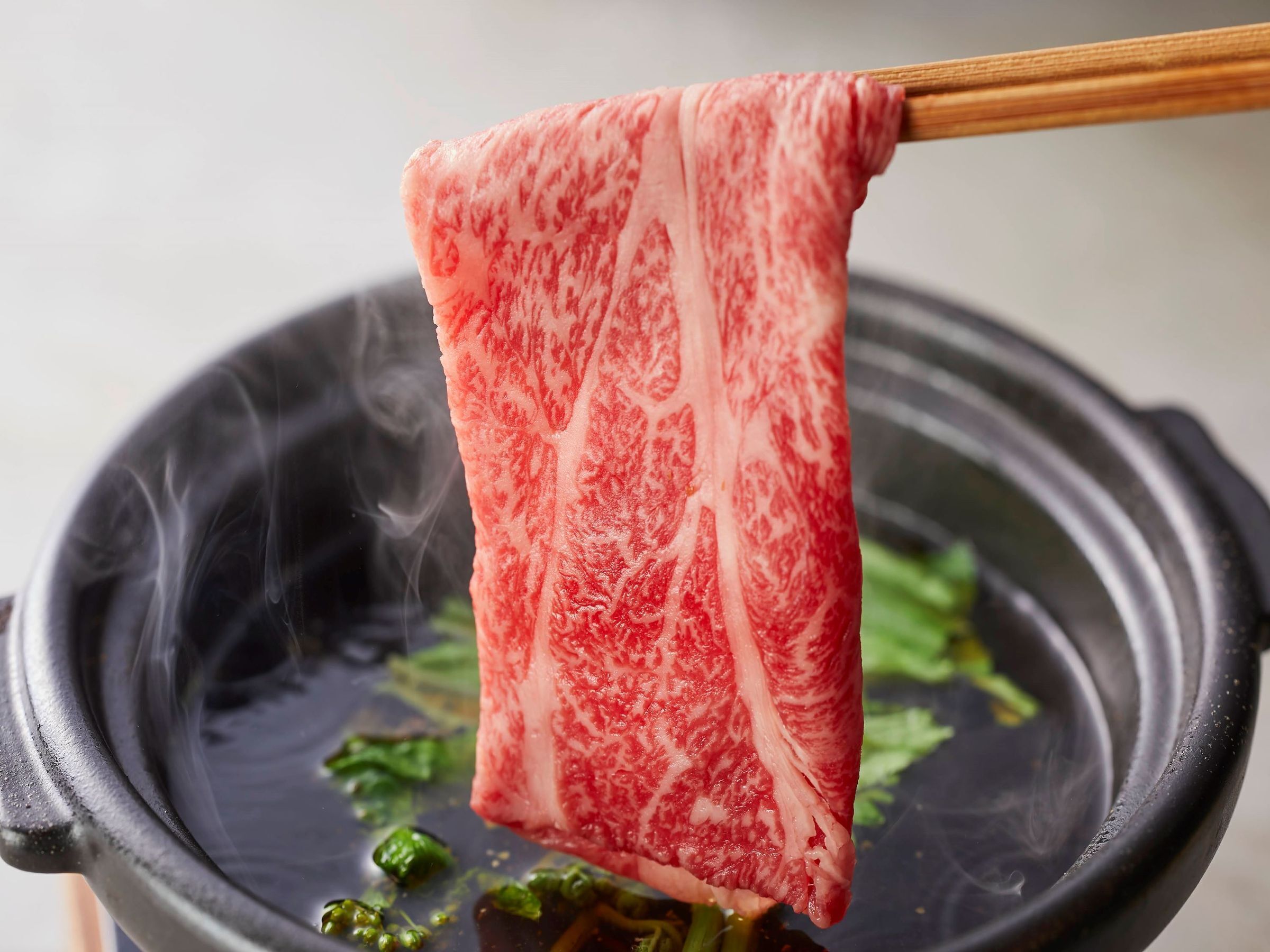 A piece of meat being held by chopsticks over a pan with steam rising, indicating it is being cooked.