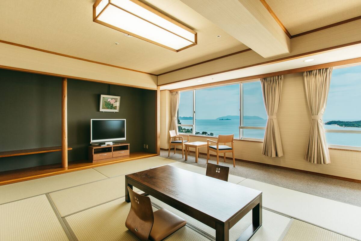 A modern room with tatami flooring, a wooden table, chairs, a TV, and a view of the sea.