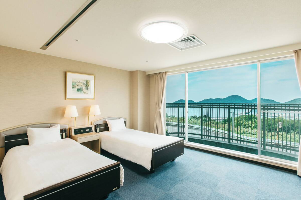 A hotel room with two beds, a large window with a view of mountains, and a ceiling light.