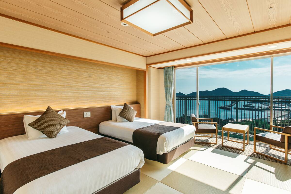 A hotel room with two beds, a balcony with a view of the sea and mountains, and a small seating area.