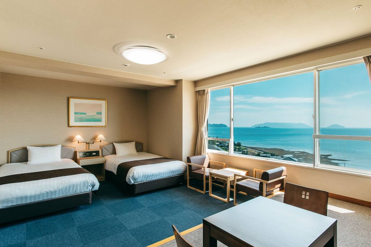 A hotel room with two beds, a desk, and a chair. There is a large window with a view of the sea and mountains.