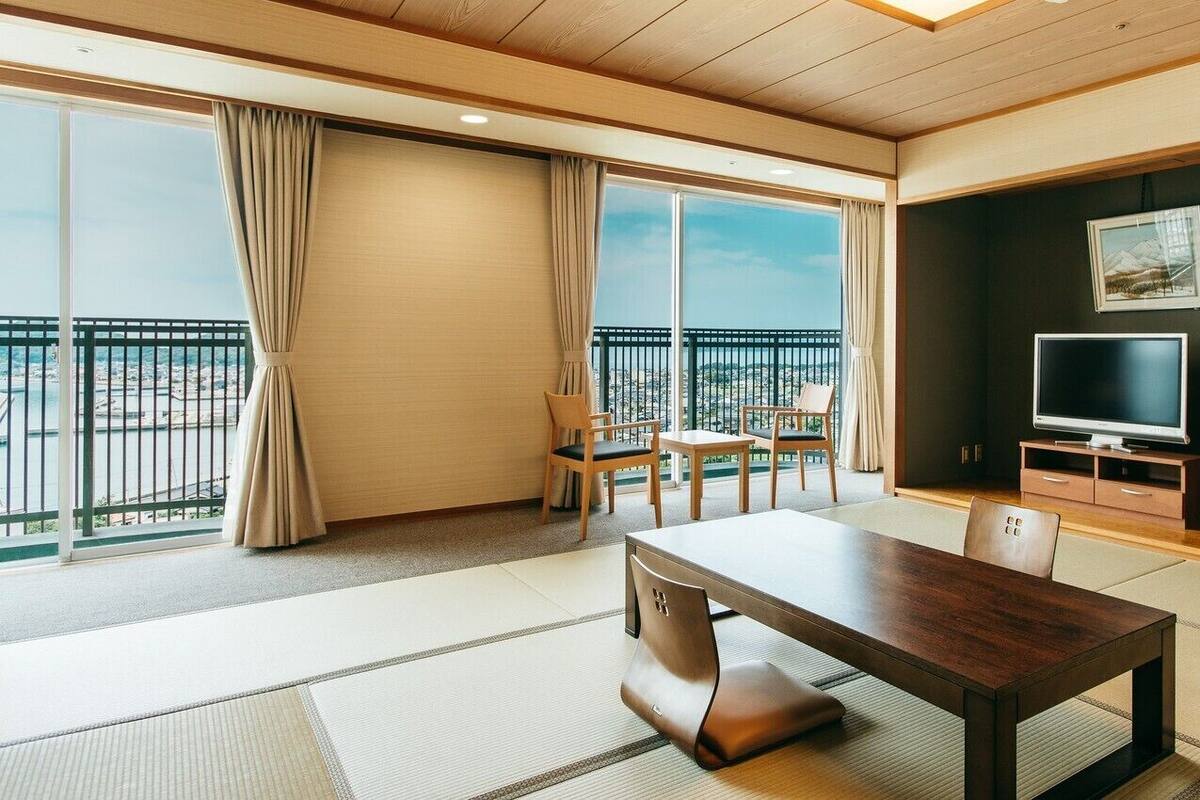 A modern hotel room with a large window, a dining table, a TV, and a balcony with a view.