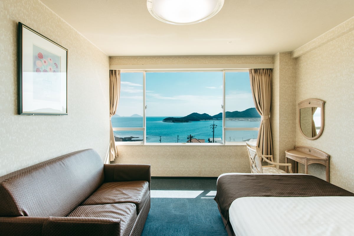 A hotel room with a large window offering a view of the sea and mountains, a bed, a sofa, a desk, and a mirror.