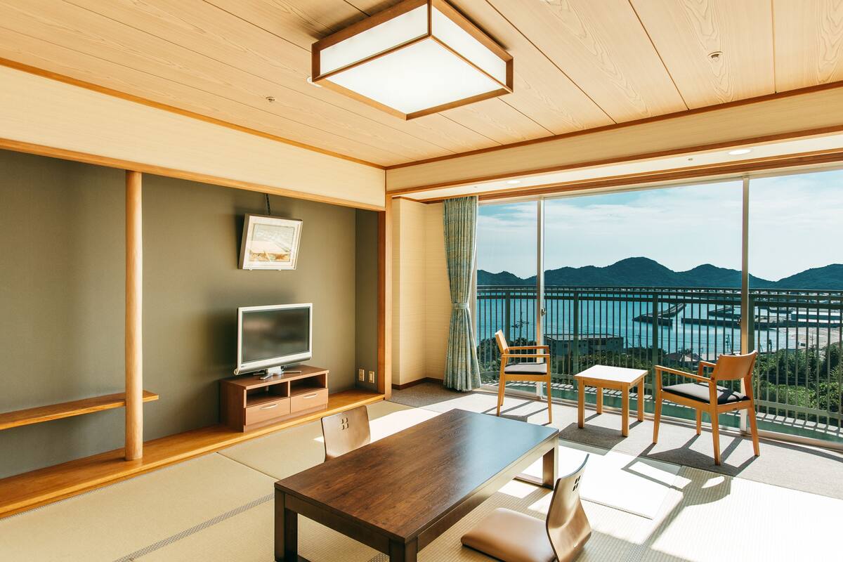 A modern hotel room with a wooden ceiling, a large window with a view of mountains and a body of water, a flat-screen TV, and a dining table with chairs.