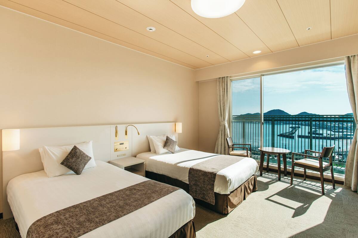A hotel room with two beds, a balcony with a view of the sea, and a small table and chair.