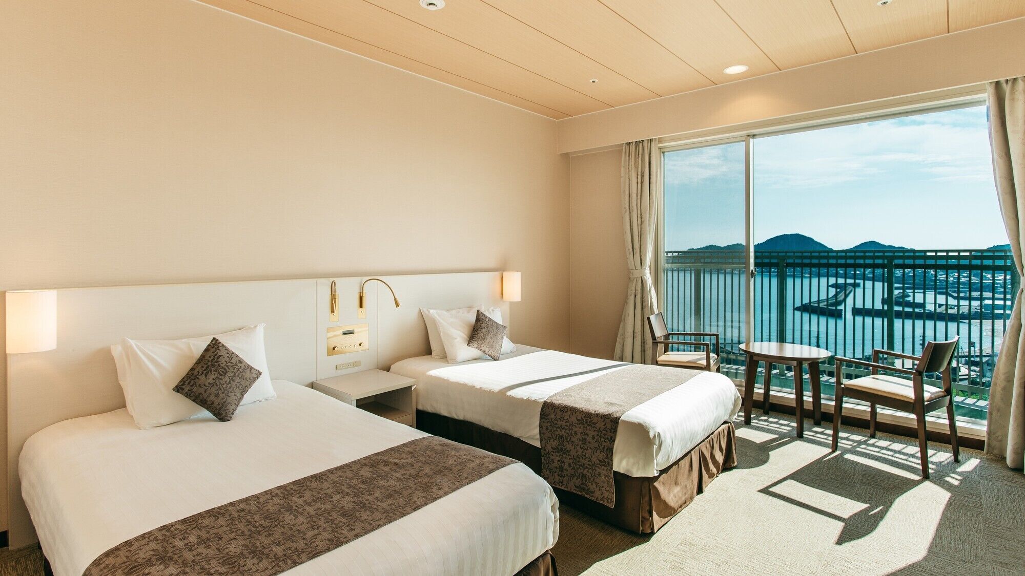 Een hotelkamer met twee bedden, een kleine tafel, een stoel en een balkon met uitzicht op zee.