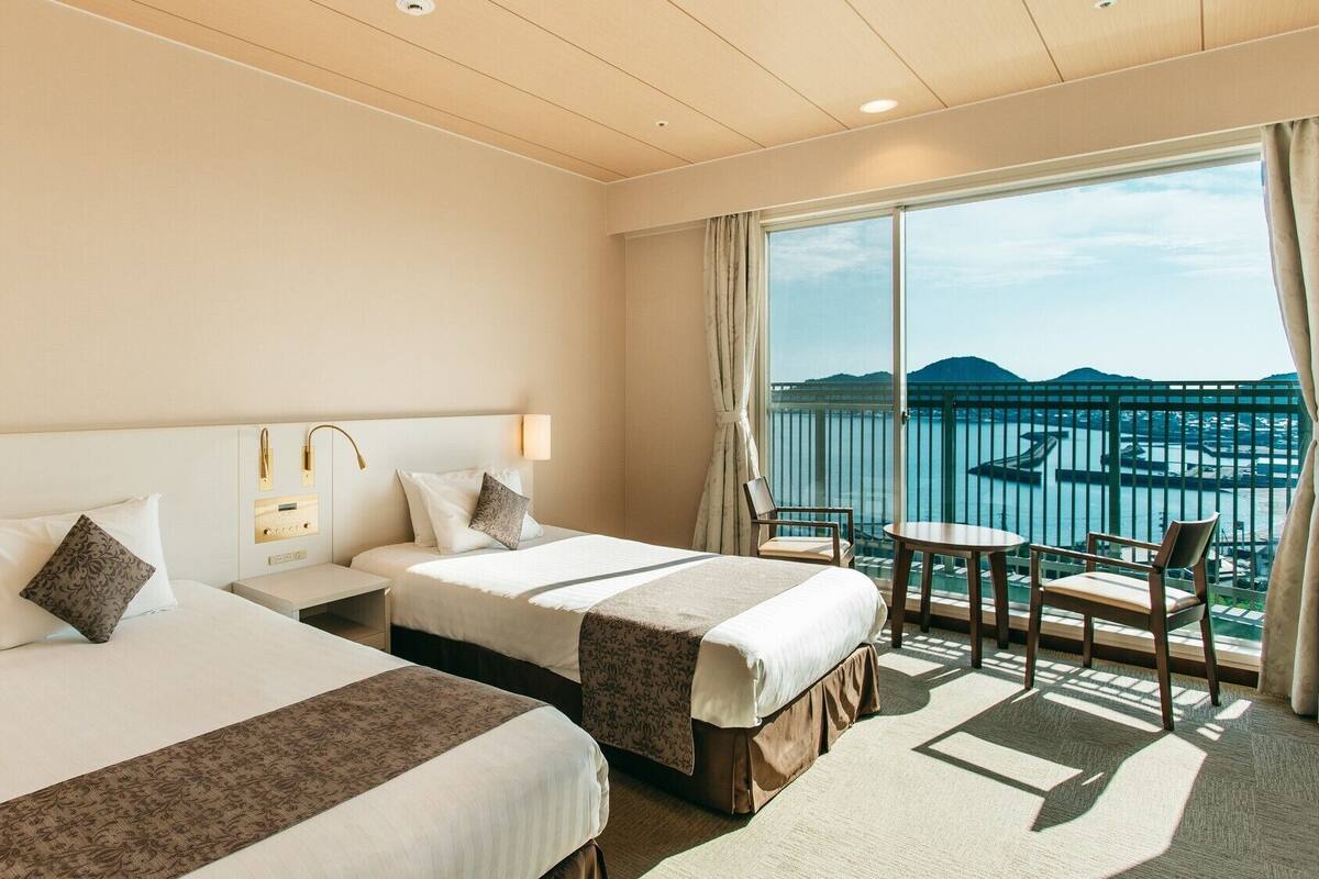 A hotel room with two beds, a small table, a chair, and a balcony with a view of the sea.