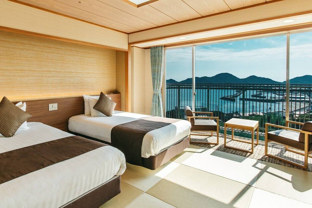 A hotel room with two beds, a balcony with a view of a body of water and mountains, and a seating area with a table and chairs.