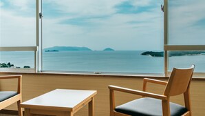 Ein Zimmer mit großem Fenster und Blick auf das Meer und entfernte Inseln. Ein Holztisch und ein Sessel mit Kissen sind vorhanden.