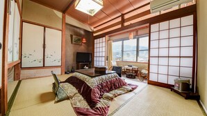 A traditional Japanese room with tatami flooring, a futon bed, sliding doors, a television, and a view of the outdoors. - Yamaguchiya Annex (Nagato)