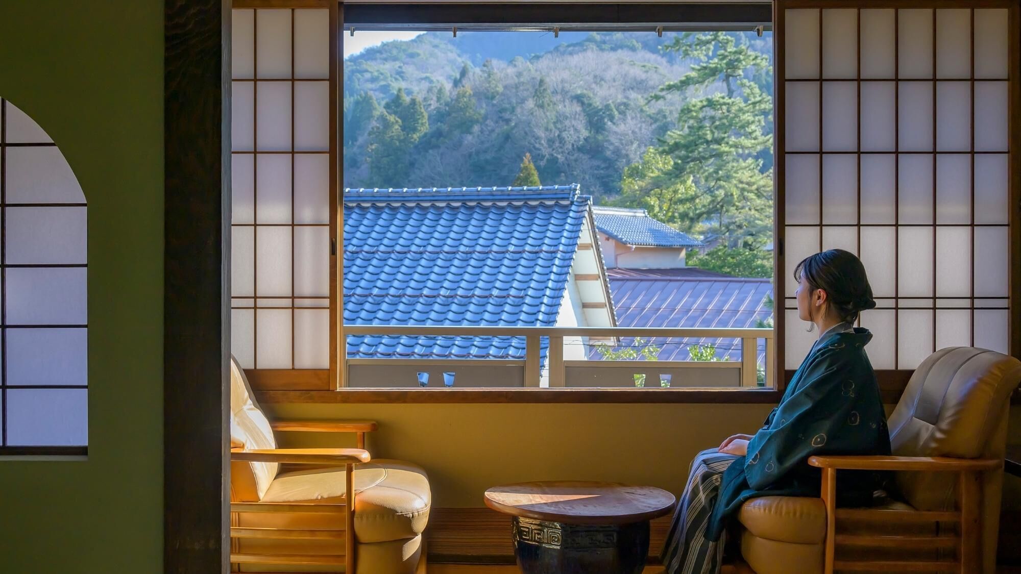 Una habitación japonesa tradicional con suelo de tatami, una mesa de madera baja y vistas a una montaña y edificios tradicionales.