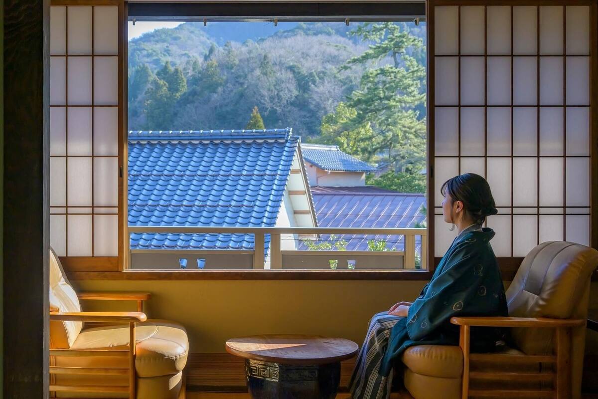 Et traditionelt japansk rum med tatami gulv, et lavt træbord og udsigt til et bjerg og traditionelle bygninger.