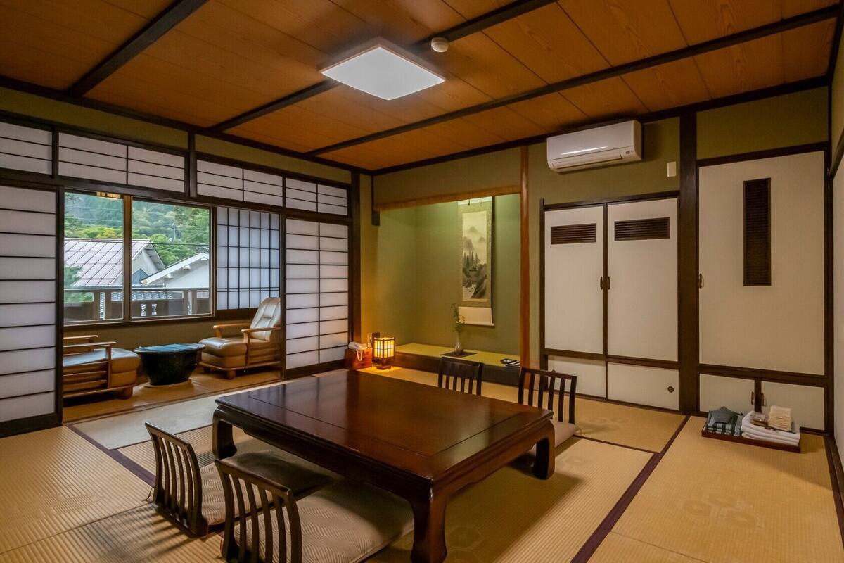 Et traditionelt japansk rum med tatamimåtter, et lavt bord, stole, en skydedør og udsigt til et boligområde.