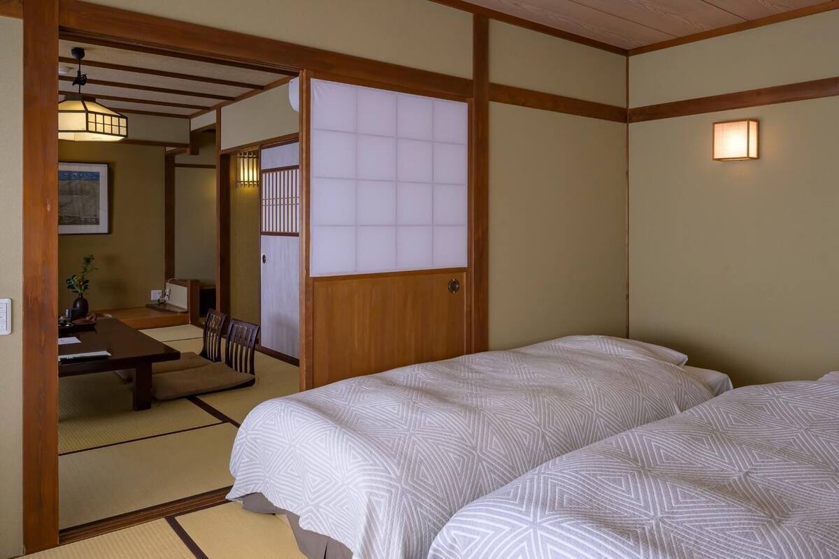 Et traditionelt japansk værelse med tatamimåtter, en lav træseng med hvidt sengetøj, en shoji-skydedør og et lille skrivebord med en stol.