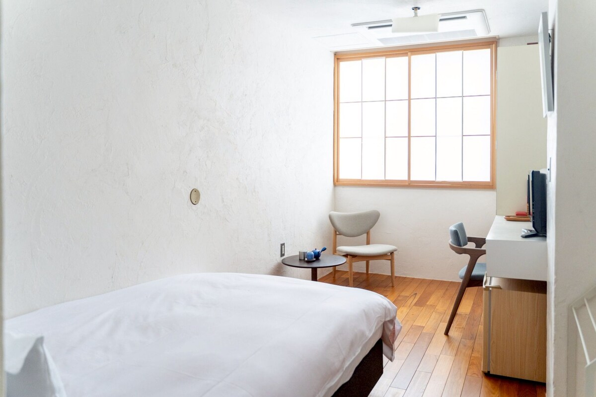 A hotel room with a bed, a small table with a chair, a desk with a chair, and a window with blinds.