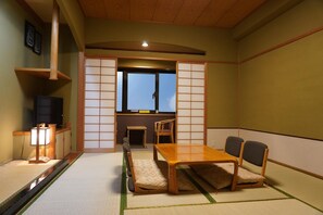 Ett traditionellt japanskt rum med tatamimattor på golvet, ett lågt bord med stolar, en tv och en skjutdörr som leder ut till ett fönster.