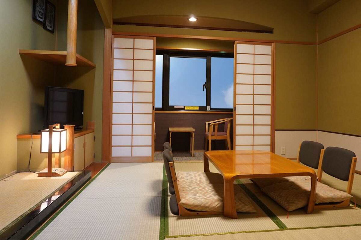 A traditional Japanese-style room with tatami flooring, a low table with cushions, a television, and sliding shoji screens.