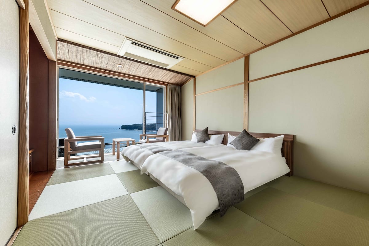 Une chambre d’hôtel avec un sol recouvert de tatamis, un grand lit, une vue sur la mer et un balcon avec une chaise.