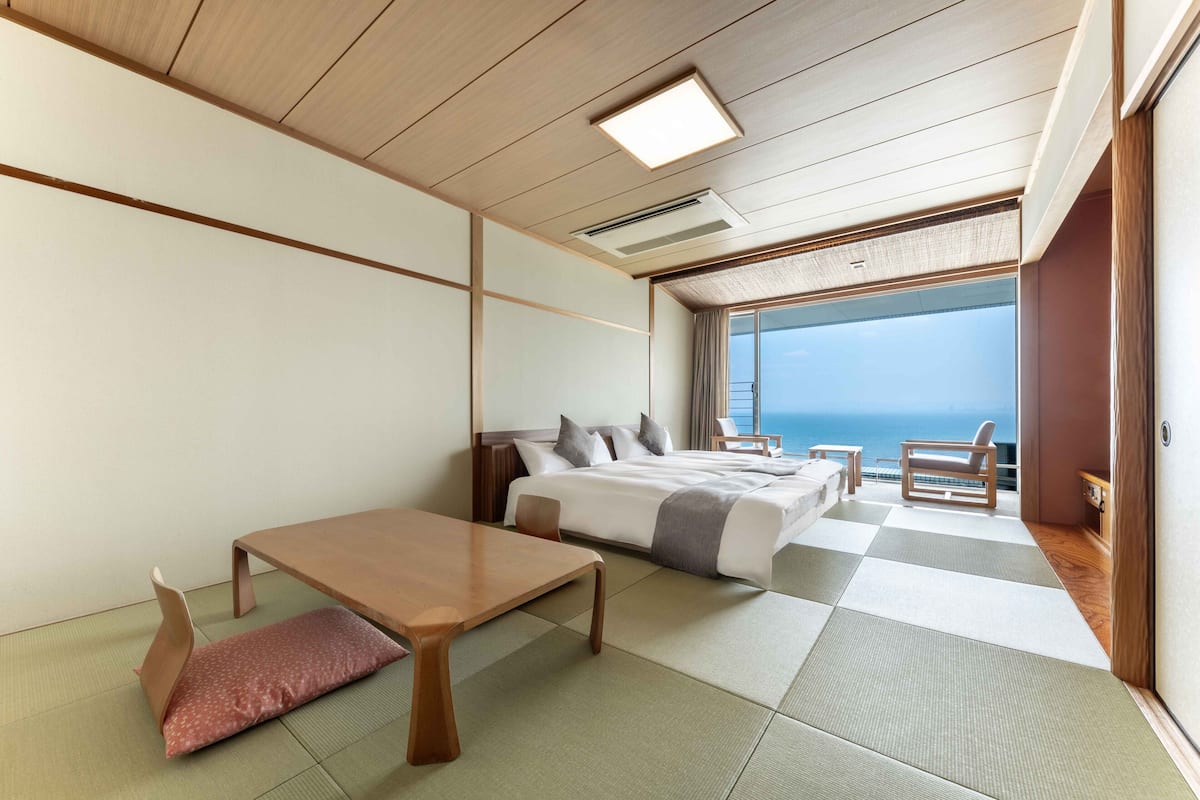 Une chambre d’hôtel avec un sol recouvert de tatamis, un lit avec des oreillers, une table en bois et une vue sur la mer.