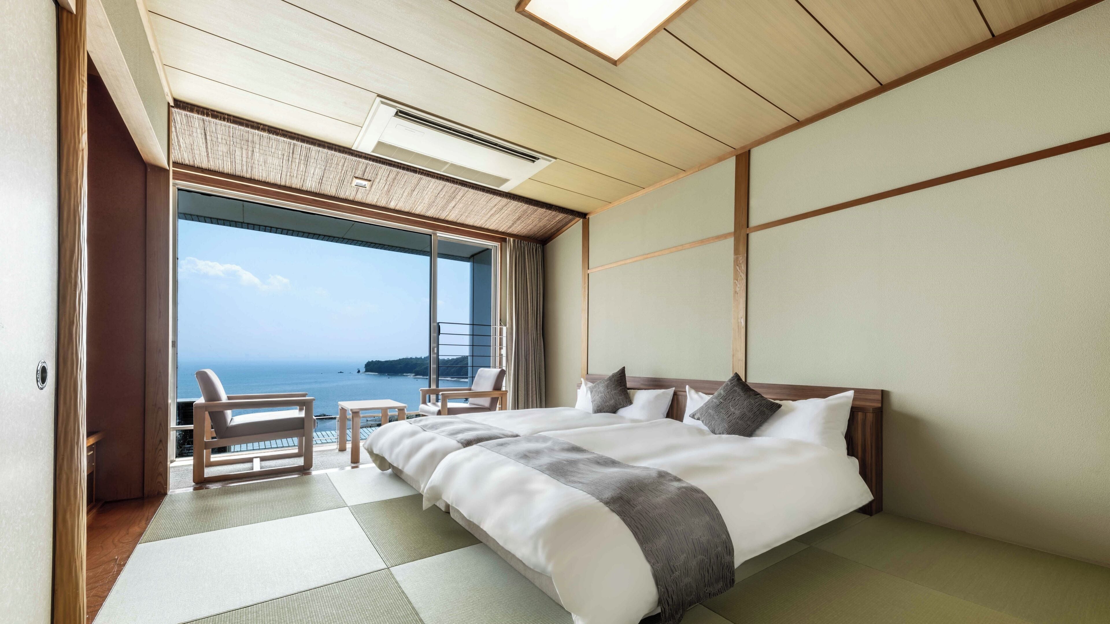 A hotel room with a large bed, a view of the sea, and a balcony with chairs.